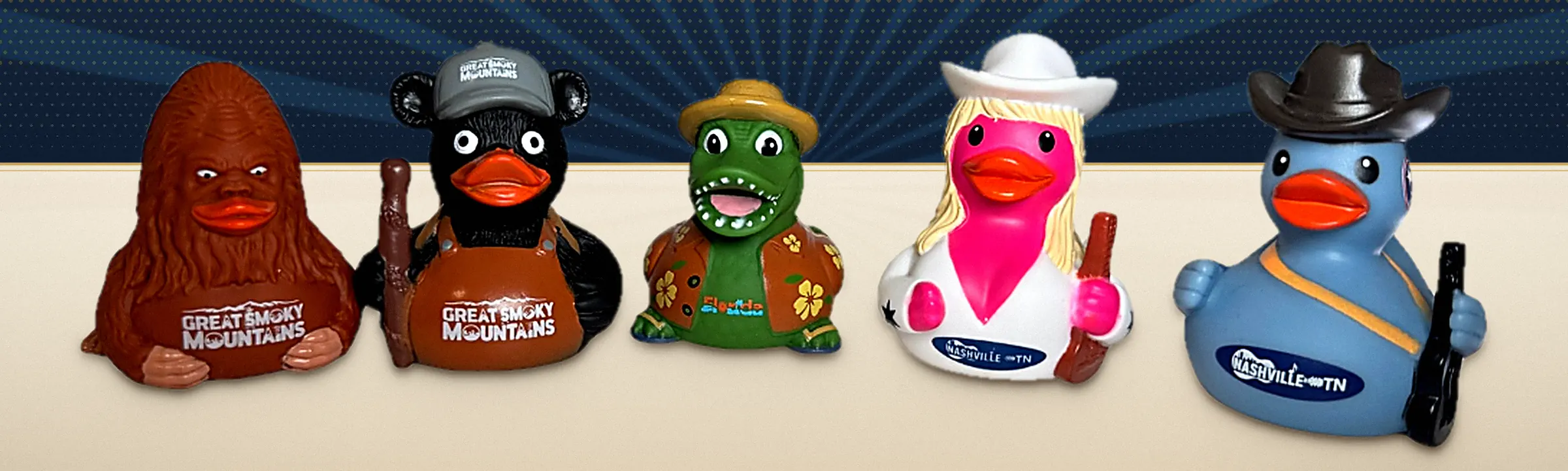 Five fully custom molded duck collectibles produced by Nash Ducks, lined up on a navy and cream vintage poster background: a brown Sasquatch, a black bear in overalls holding a club, a green alligator in a Hawaiian shirt and straw hat, a blonde country singer in a white cowboy hat holding a guitar, and a blue cowboy duck in a black hat holding a guitar
